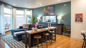 A modern living room with a dark blue sofa, a wooden coffee table, and a city view through large windows. - ★HostWise Stays★ Rooftop Deck | Garage | Balcony Views | Upscale! (Pittsburgh)