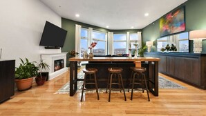 A modern kitchen with a central island, bar stools, a flat-screen TV, and large windows offering a city view. - ★HostWise Stays★ Rooftop Deck | Garage | Balcony Views | Upscale! (Pittsburgh)