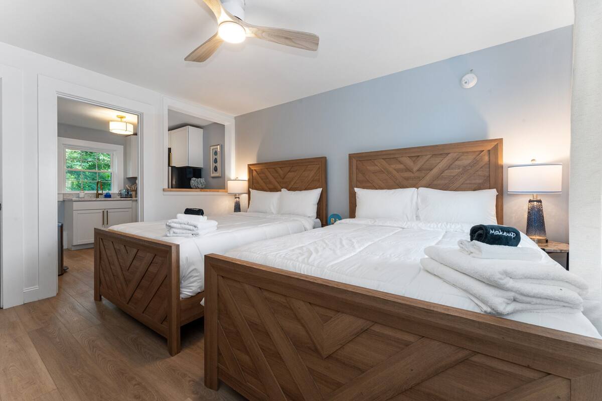 A hotel room with two beds, a ceiling fan, a nightstand with a lamp, and a view of the kitchen and window.