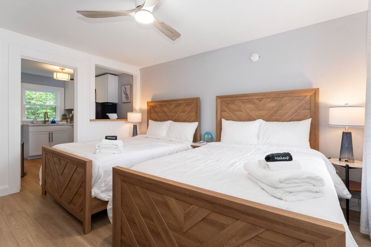 A hotel room with two beds, wooden headboards, white bedding, a ceiling fan, a small table with a lamp, and a view of the outside.