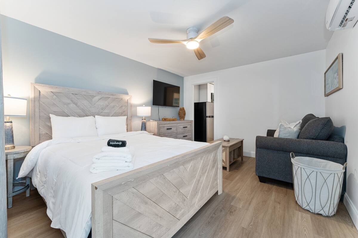 A bedroom with a bed, a ceiling fan, a TV, a grey sofa, and a trash bin.
