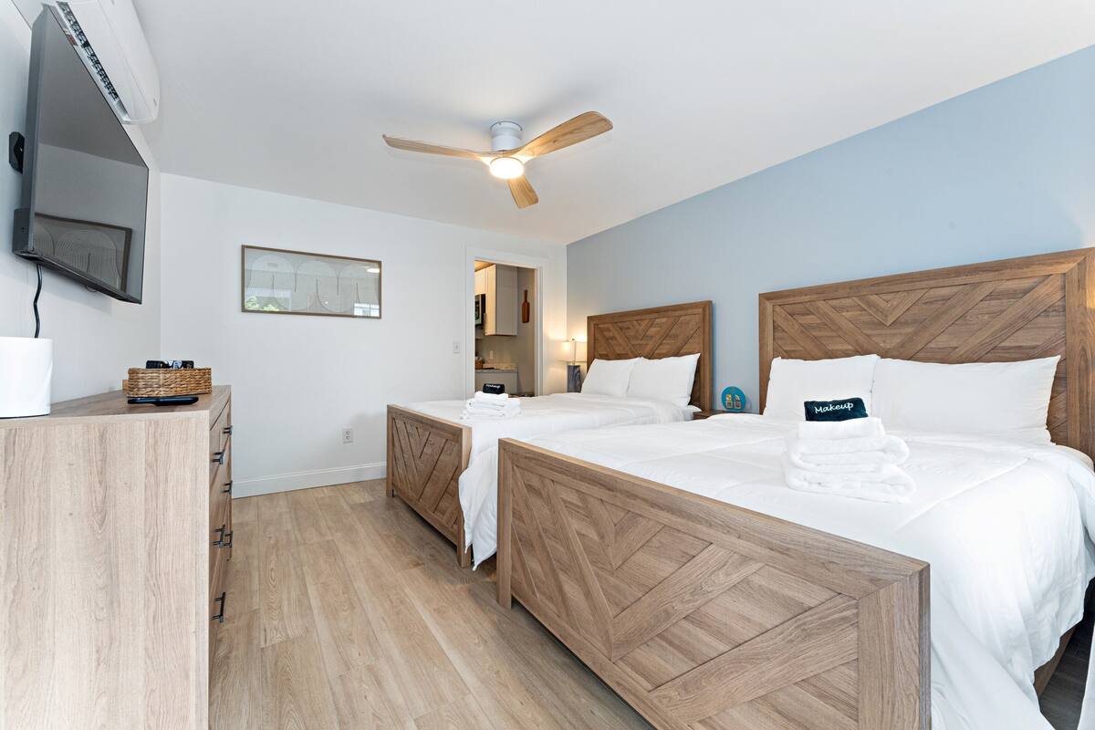 A hotel room with two wooden beds, a ceiling fan, a flat-screen TV, a wooden dresser, and a framed picture on the wall.