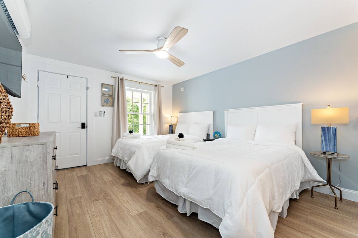 A bedroom with two beds, a ceiling fan, a bedside table with a lamp, and a window with curtains.