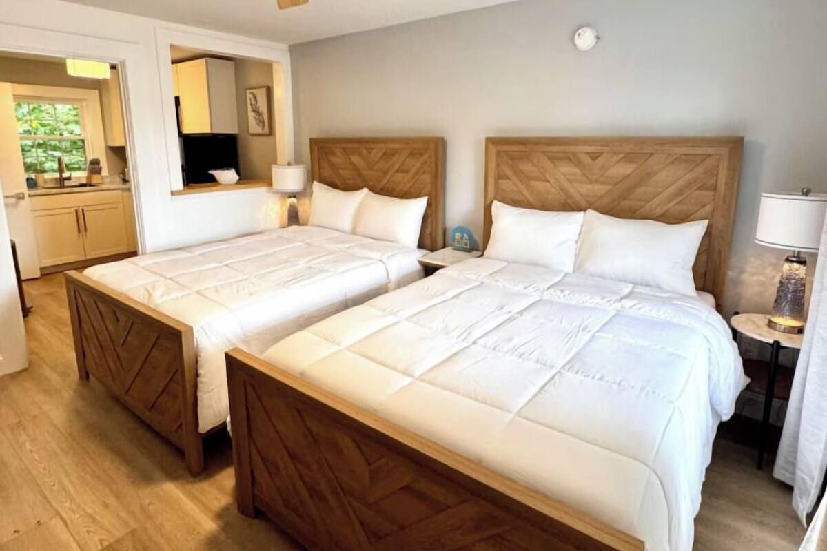 A hotel room with two beds, wooden headboards, a ceiling fan, a bedside table with a lamp, and a view of the kitchen area.
