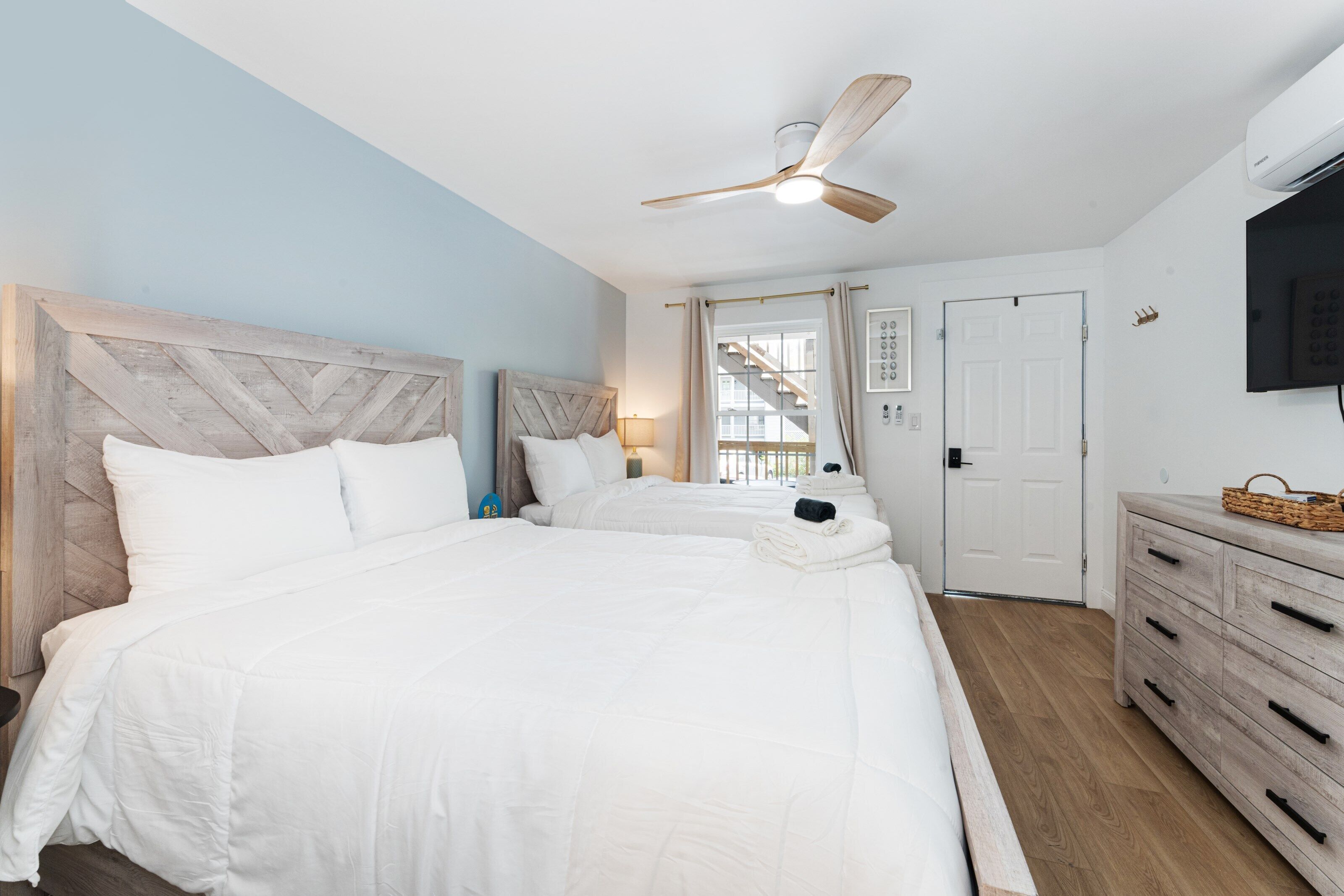 Habitación de hotel con una cama grande, ventilador de techo, televisión, cómoda de madera y una escalera visible a través de la ventana.