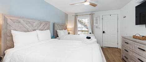 Habitación de hotel con una cama grande, ventilador de techo, televisión, cómoda de madera y una escalera visible a través de la ventana.