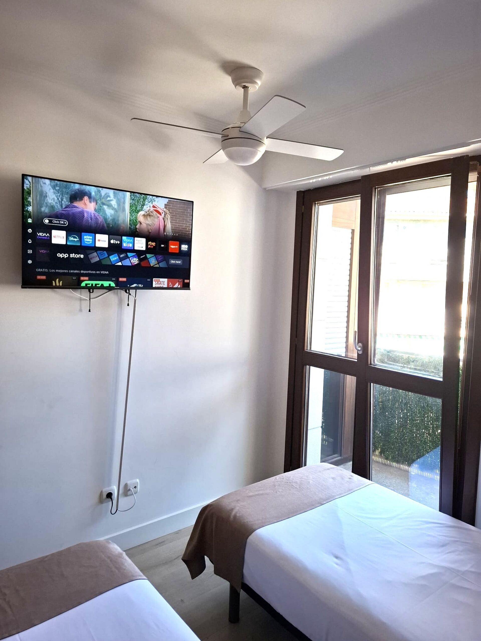 Habitación de hotel con televisor de pantalla plana montado en la pared que muestra varios canales, ventilador de techo y un amplio ventanal con vistas al exterior.