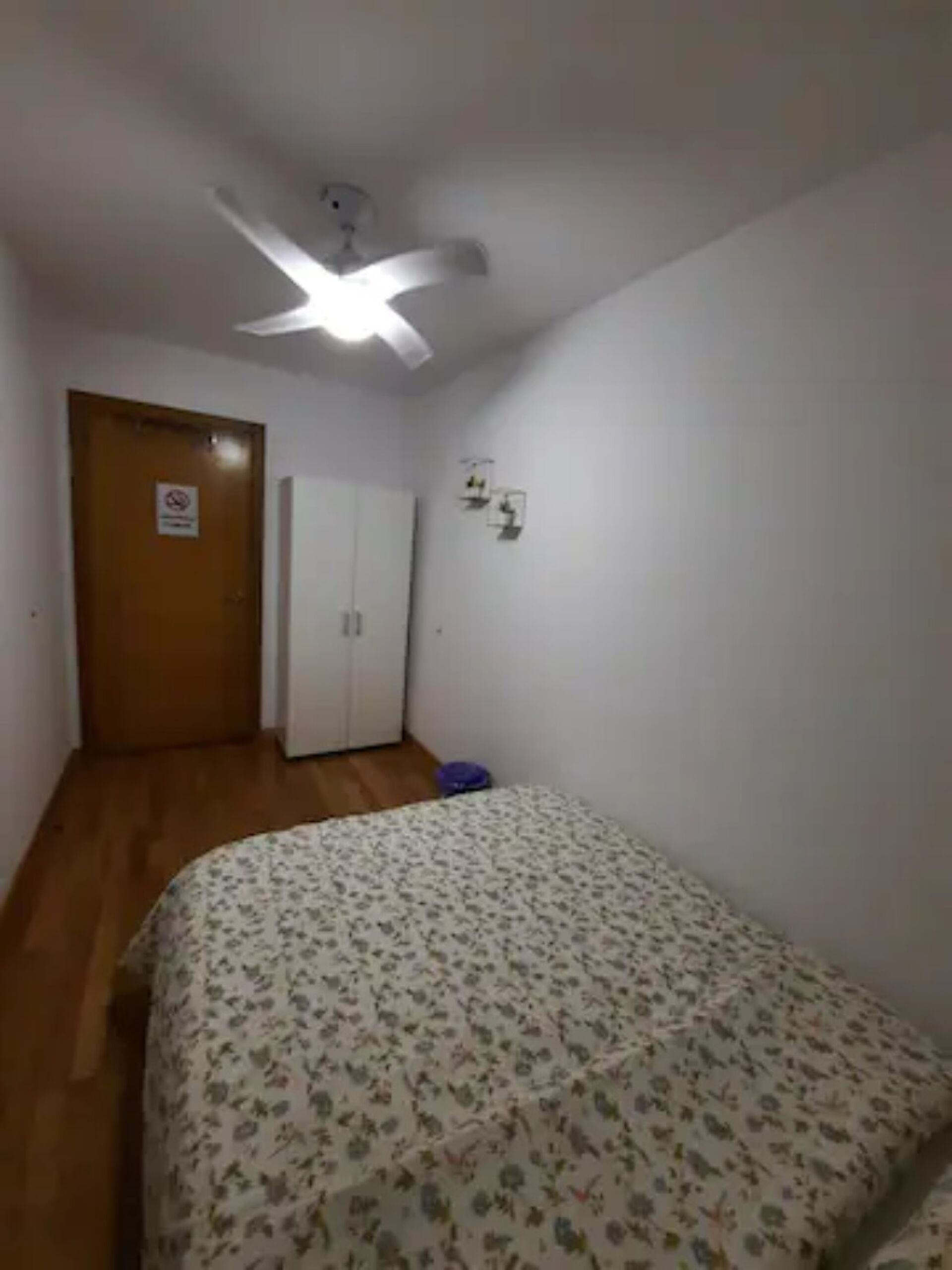 A bedroom with a bed, a ceiling fan, a wardrobe, and a door with a sign.