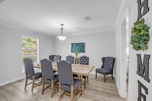 Un coin repas comprenant une table en bois, six chaises rembourrées, un lustre et une fenêtre avec des stores.