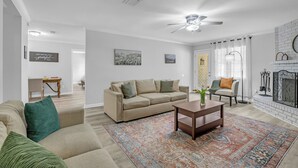 Olohuoneessa on beige sohva, sohvapöytä, kuviollinen matto, takka ja kattotuuletin.