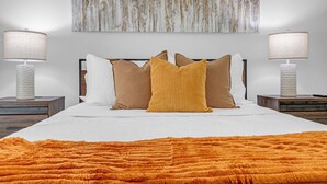 Un lit bien fait, recouvert d’une couverture blanche et orange, avec deux lampes de chevet et un grand tableau abstrait accroché au mur.