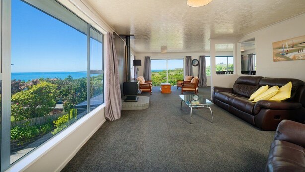 Ein geräumiges Wohnzimmer mit großem Fenster und Meer- und Grünblick, ein braunes Ledersofa, ein Glastisch und eine moderne Pendelleuchte.