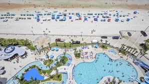 Ovenfra set et strandresort med et stort poolområde, strandparasoller og en sandet kystlinje.