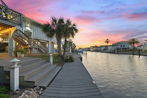 Une propriété située au bord de l’eau, dotée d’une terrasse en bois, d’un escalier menant à un bâtiment, de palmiers et offrant une vue sur le coucher de soleil sur l’eau.