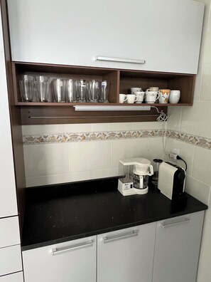 Una cocina compacta con encimera negra, cafetera y estante con copas y tazas.