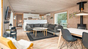 Een moderne woonkamer met een bank, salontafel en stoelen, een keuken op de achtergrond, houten wanden en een groot raam.