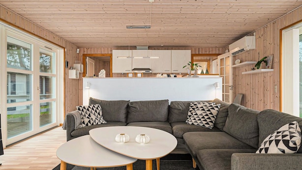 Een moderne woonkamer met een grijze modulaire bank, twee witte ronde tafels, en een keuken met houten kasten en een wit aanrecht.