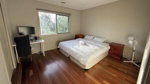 Un dormitorio con cama, un escritorio con ordenador, una ventana y un ventilador de pie.