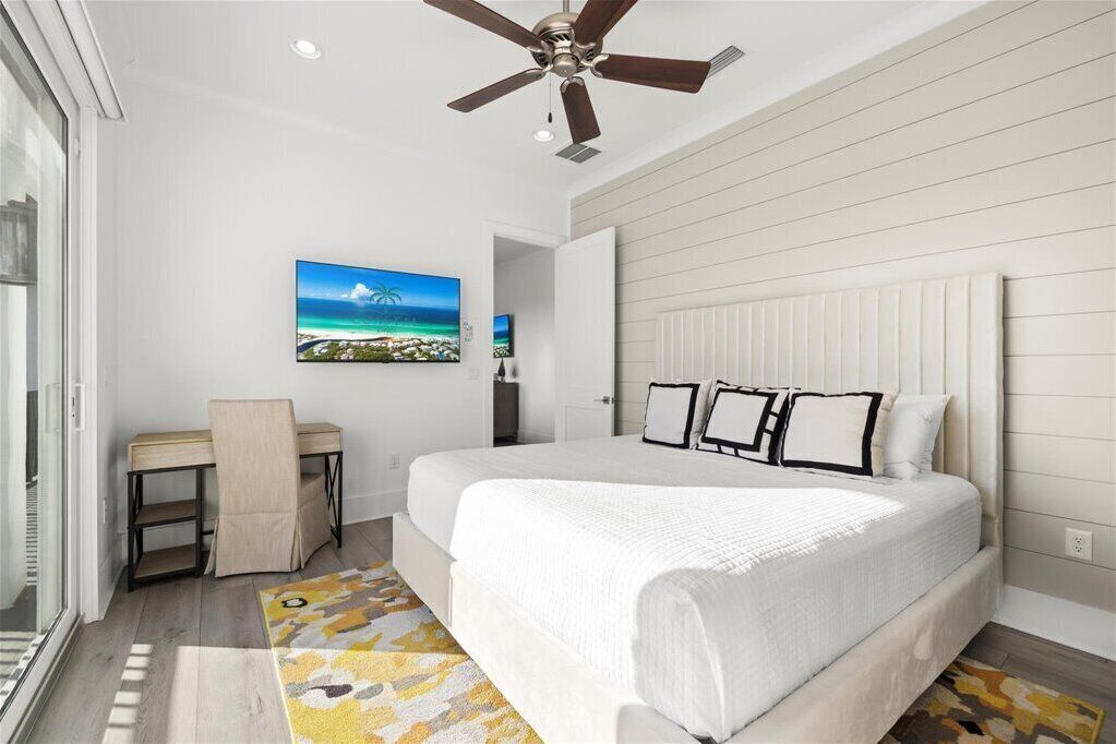 A bedroom with a bed, a ceiling fan, a desk, a chair, a window, and a painting on the wall.