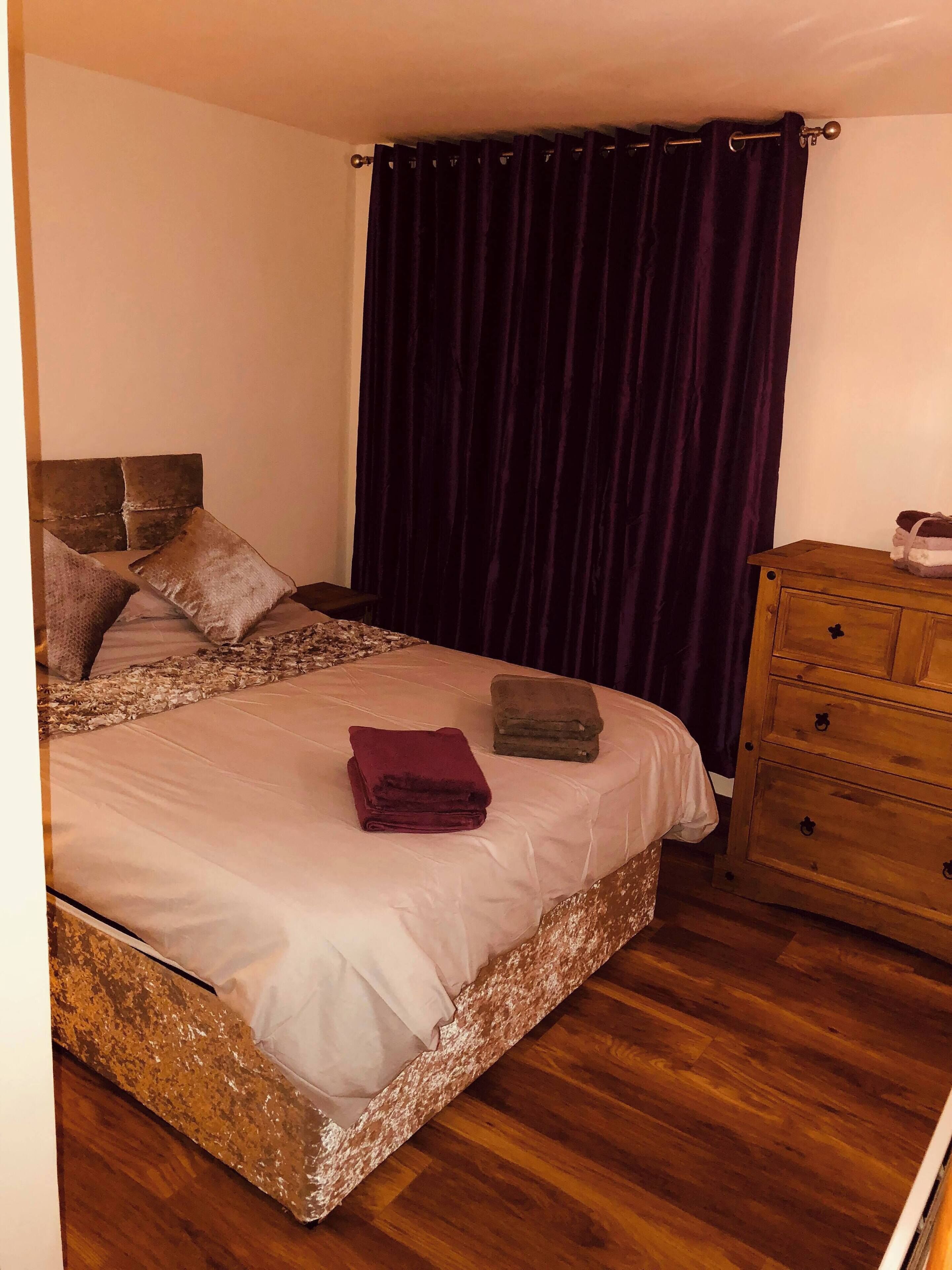 Een slaapkamer met een bed, kussens, een houten dressoir en een donker gordijn.