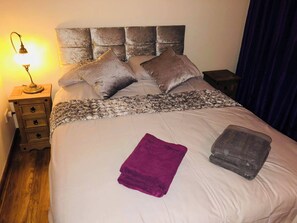 Un lit bien fait, avec une tête de lit, une lampe de chevet, une table de chevet en bois dotée de tiroirs, et une serviette violette posée sur le lit.