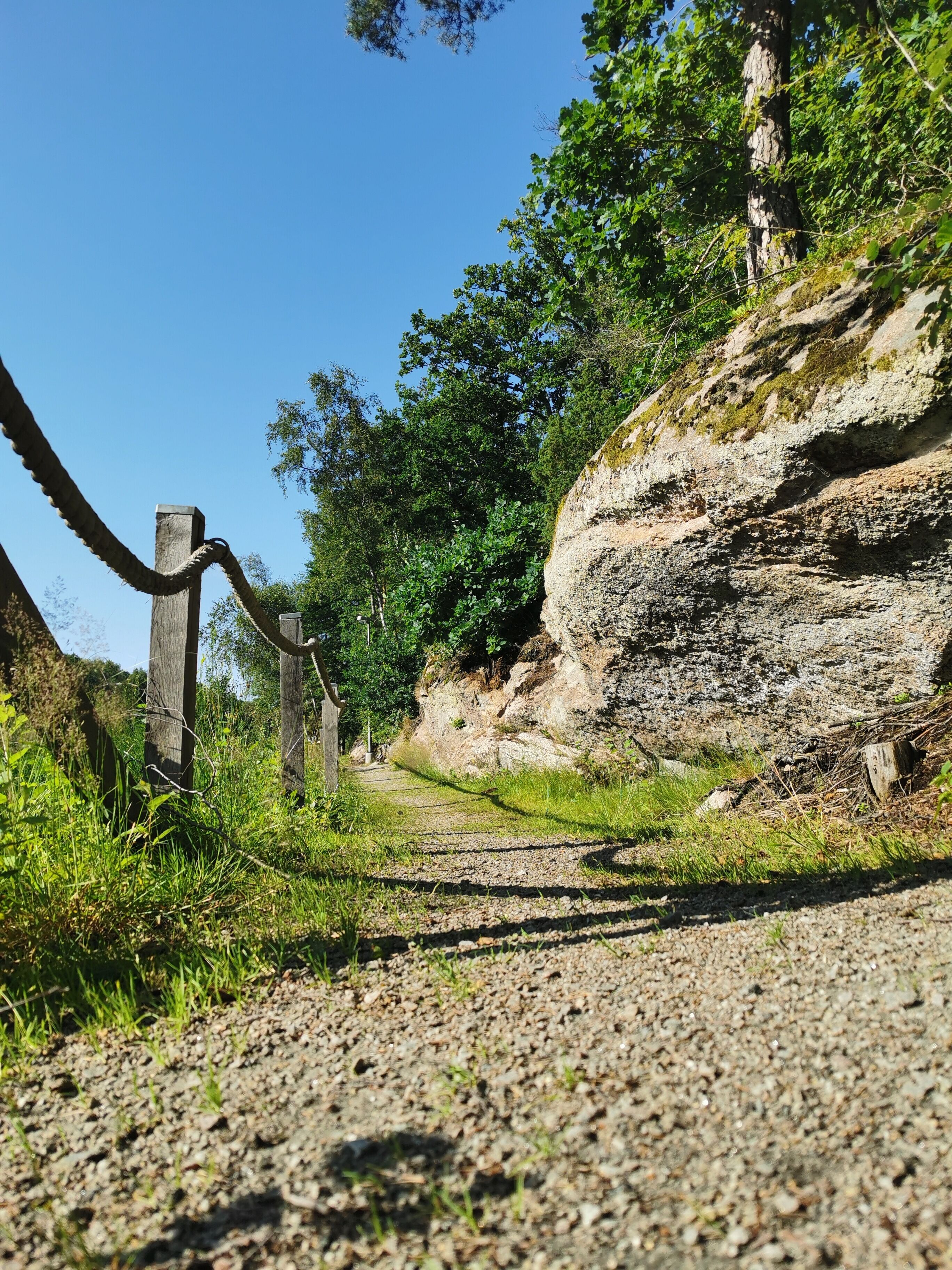 Un sentier de gravier traverse un paysage naturel, bordé d’une clôture en bois et présentant un terrain rocheux.