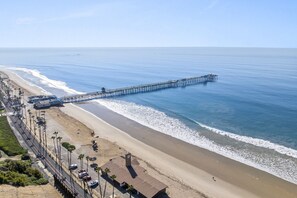 Una playa con un muelle largo que se adentra en el océano, una orilla arenosa y una carretera con coches aparcados y palmeras.