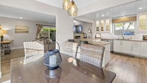 A modern kitchen with white cabinets, a large island, and a dining area with wicker chairs and a glass table. - Hideaway 4Plex! Walk to the beach, Downtown, AC! (San Clemente)