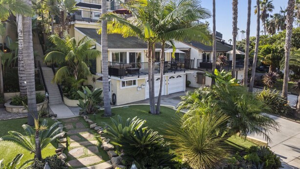 Una propiedad residencial con una casa de dos pisos, un balcón con mobiliario exterior y un jardín bien cuidado con palmeras y otras plantas tropicales.