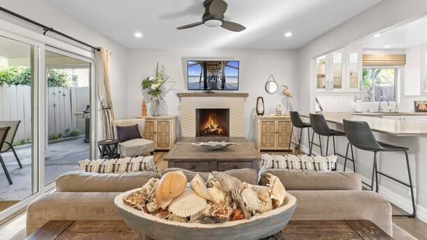 A spacious living room with a fireplace, comfortable seating, and a large window overlooking a garden. - Hideaway A- Walk to the beach, Downtown, AC! (San Clemente)
