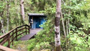 Eine Holzbrücke führt zu einer kleinen, blauen Holzhütte in einem bewaldeten Gebiet.