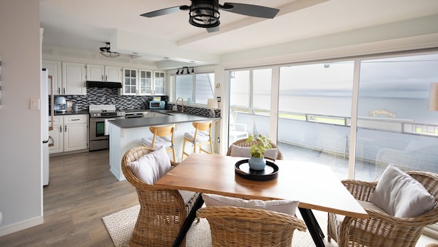 Cozinha moderna com armários brancos, painel de backsplash preto e branco, e uma ilha grande. Há uma área de refeições com cadeiras de vime e uma mesa de madeira.