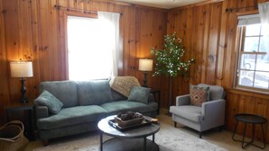 Een woonkamer met een bank, fauteuil, en salontafel, houten wanden, en een raam met gordijnen.