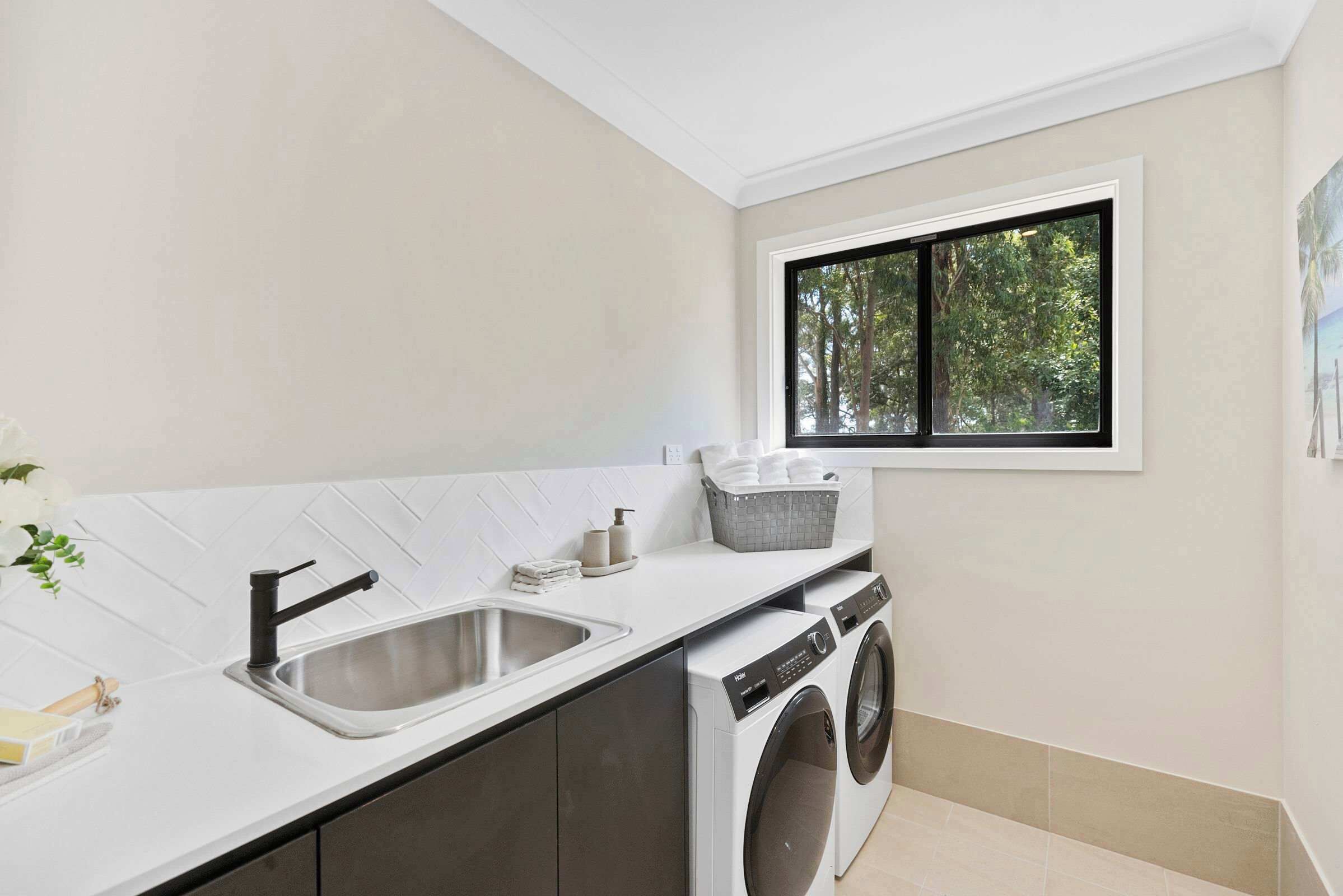 A modern laundry room with a sink, washing machine, and a window with a view of trees.