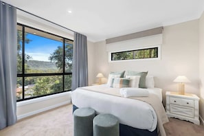 A bedroom with a large window, a bed with pillows, a nightstand, and two stools. - Ruby Lodge by Coast Hosting (Green Point)