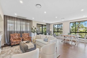 A spacious living room with a large window, a sofa, chairs, and a dining area with a table and chairs. - Sun-Drenched. Brand-New Home. Sleeps 11 (Green Point)