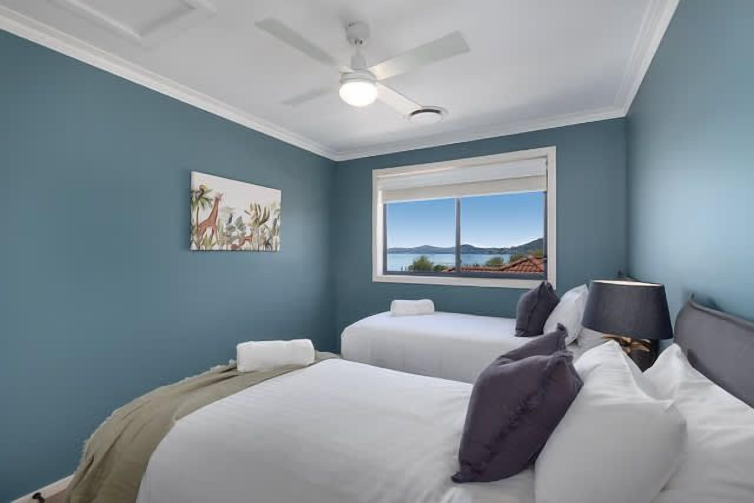 Une chambre avec deux lits, un ventilateur de plafond, une fenêtre donnant sur un beau paysage et un tableau accroché au mur.