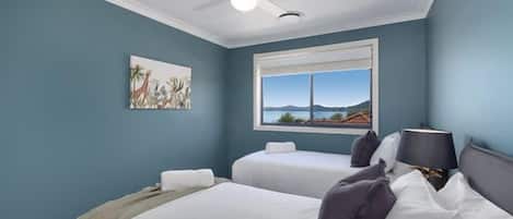 A bedroom with two beds, a ceiling fan, a window with a view, and a painting on the wall.