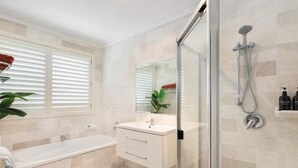 A modern bathroom with a glass-enclosed shower, a white vanity, and a large window with blinds. - Private Jetty. Waterfront. Sleeps 10 (Koolewong)
