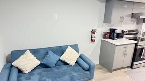 A modern living room with a blue sofa, two white cushions, a coffee maker, and a kitchen area with grey cabinets and a stove. - Zillion’s Bliss - Basement Suite (Calgary)