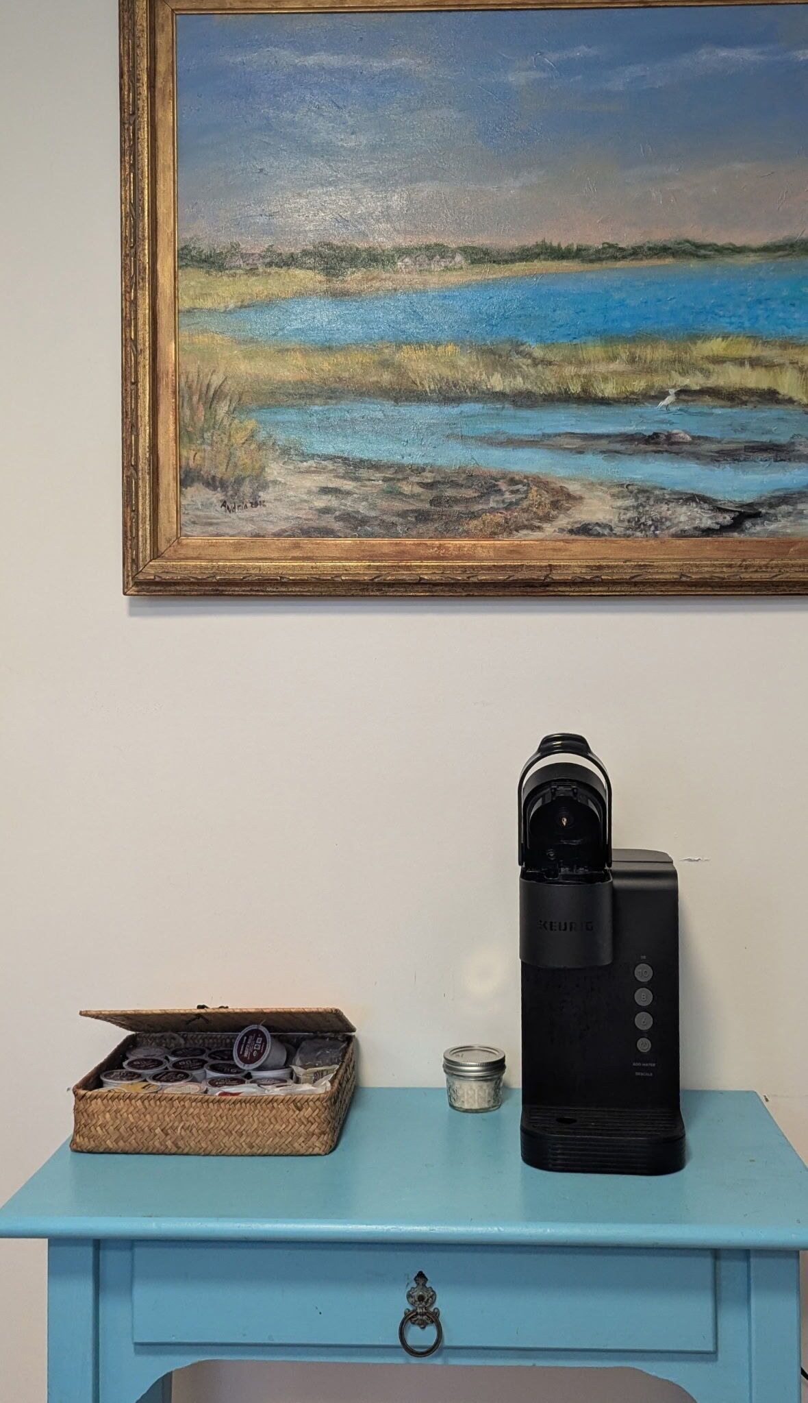 Un cuadro de un paisaje con agua y césped colgado en una pared. Debajo del cuadro, hay un mueble azul con una cafetera y una cesta de bolsitas de té.