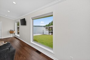 A room with a large window, a flat-screen TV, a wooden floor, and a basket on a small table. - Lakefront Home with Hot Tub, Private Dock & Ramp (Hot Springs)