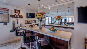 A modern kitchen with a bar area, featuring a large mirror, pendant lights, and a vase with flowers.