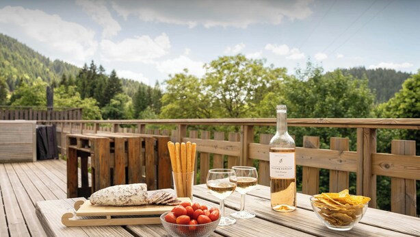 A wooden deck with a table set for a meal, including a bottle of wine, glasses, breadsticks, a loaf of bread, and a bowl of tomatoes. - La Ferme Geromoise - Gathering and exploring the Vosges - OVO Network (Gérardmer)