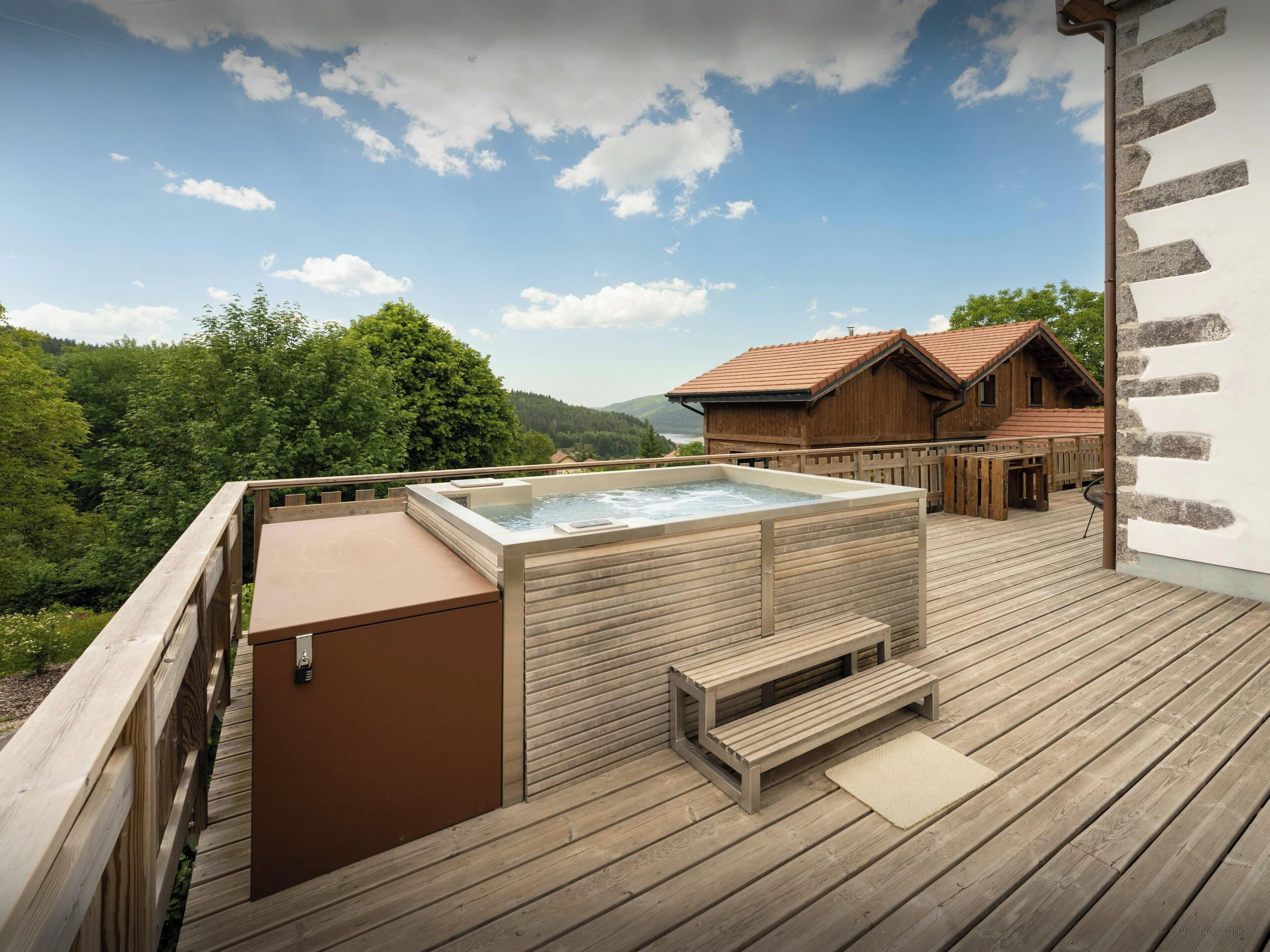 A wooden deck with a hot tub, wooden benches, and a view of a mountain and forest.
