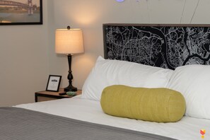 A neatly made bed with white linens and a single mustard-colored pillow, a bedside lamp, and a framed picture on a nightstand. - Stylish Townhouse with Game Room, King Suite (Cincinnati)