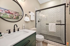 A bathroom with a white sink, black faucets, a toilet, and a glass-enclosed shower with white tiles. A painting of a cityscape is on the wall. - Stylish Townhouse with Game Room, King Suite (Cincinnati)
