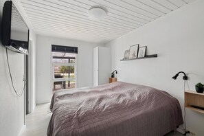 Ein modernes Schlafzimmer mit einem großen Bett, einem an der Wand montierten Fernseher, einem Schreibtisch mit Stuhl und einem Fenster mit Jalousien.