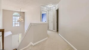 A hallway with a carpeted floor, a white wall, a door, and a window with a chandelier. - Spacious Retreat in Prime Location — Comfort, Style & Convenience” (Owings Mills)