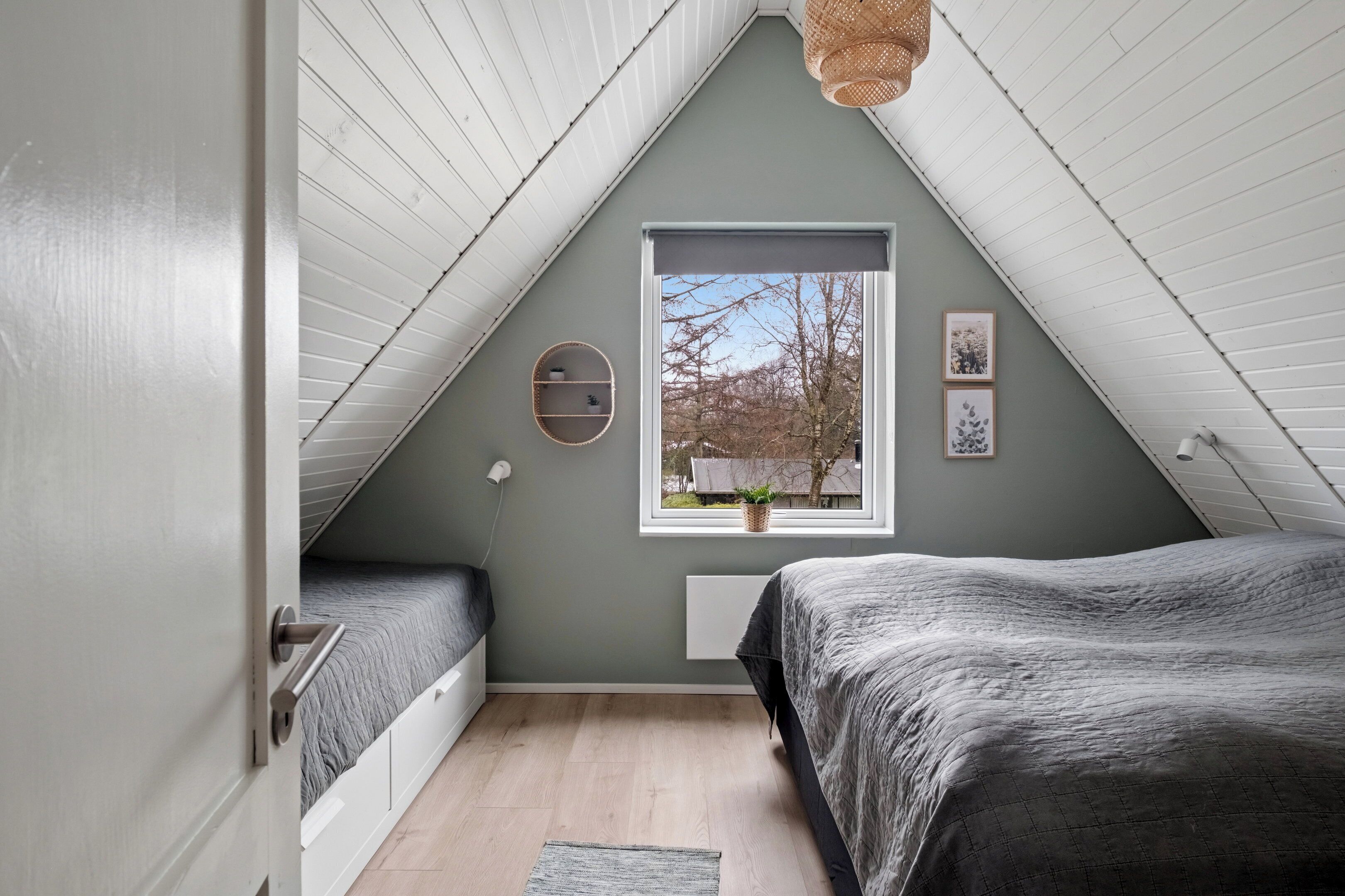 Quarto com teto inclinado, janela com vista para árvores, cama com colcha cinza e uma pequena estante.